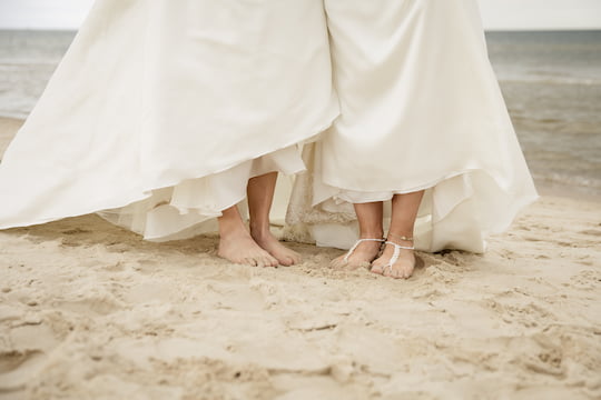 Zwei Personen (sichtbar von der Hüfte abwärts) in weissen Kleidern stehen nebeneinader barfuss am Strand vor dem Meer